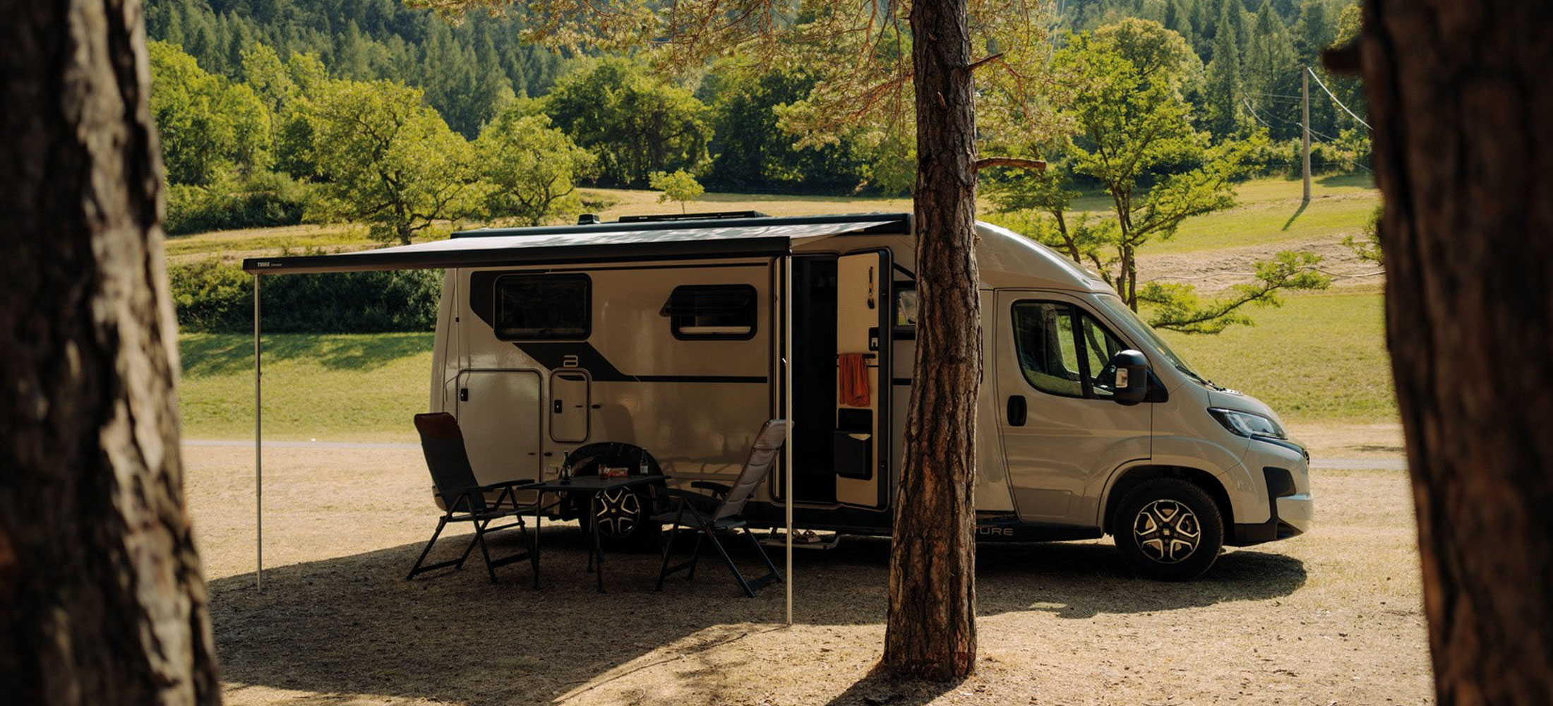 Wohnmobil auf einem naturnahen Stellplatz im Wald mit ausgefahrener Markise, geöffneter Seitentür und Campingstühlen.