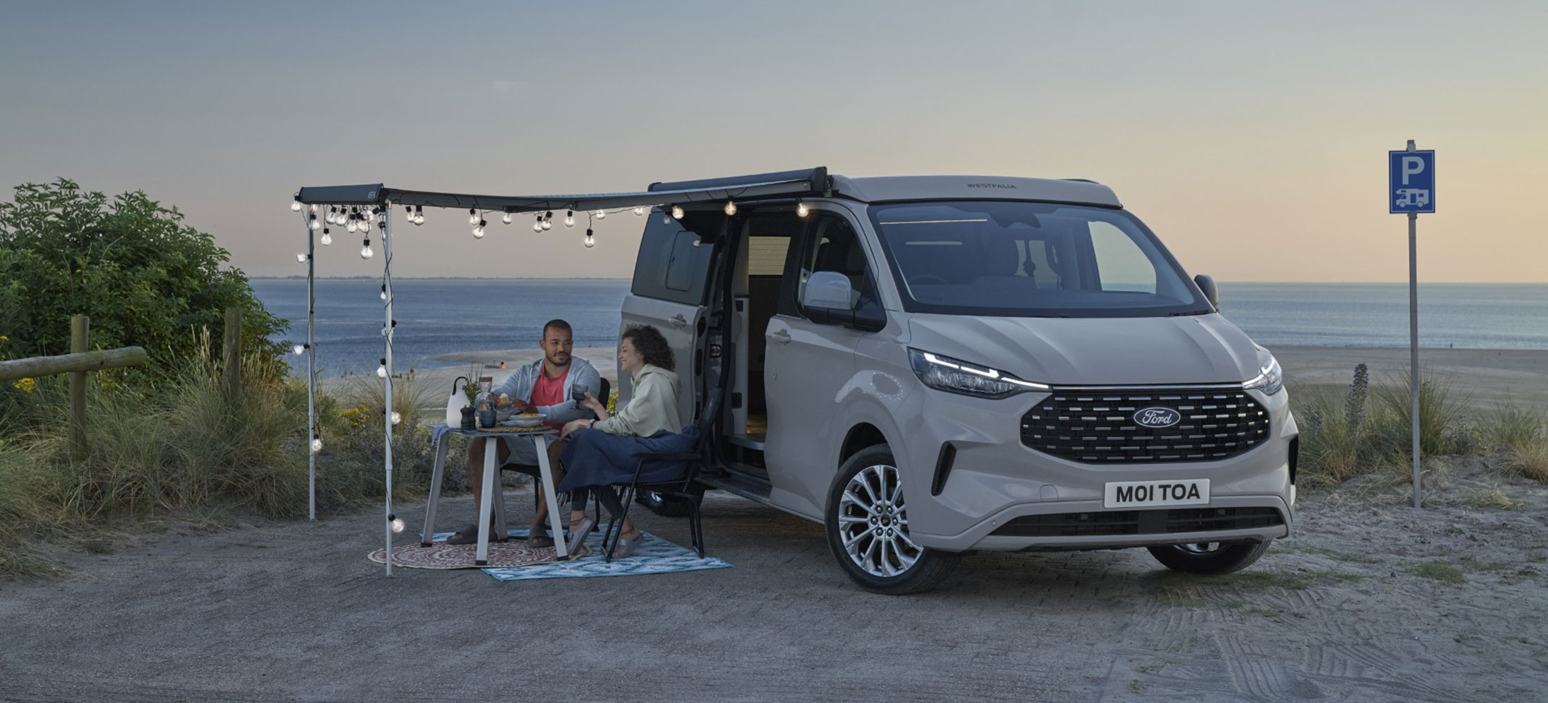 Ford Nugget als L2 in Silber vor einem Strand mit zwei Personen die unter der Markise sitzen