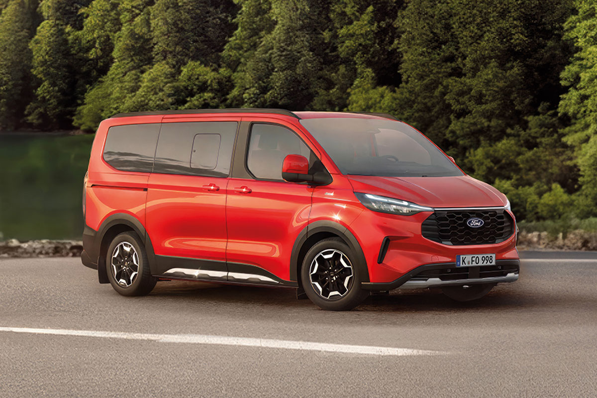 Roter Ford Tourneo Custom fährt auf einer Straße entlang eines Sees mit Waldlandschaft im Hintergrund.