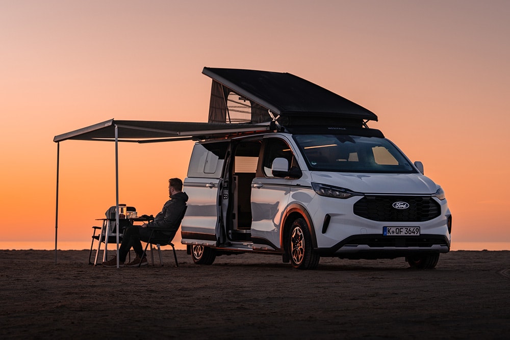 Ford Campervan mit geöffnetem Aufstelldach und ausgefahrener Markise auf einem Campingplatz bei Sonnenuntergang.
