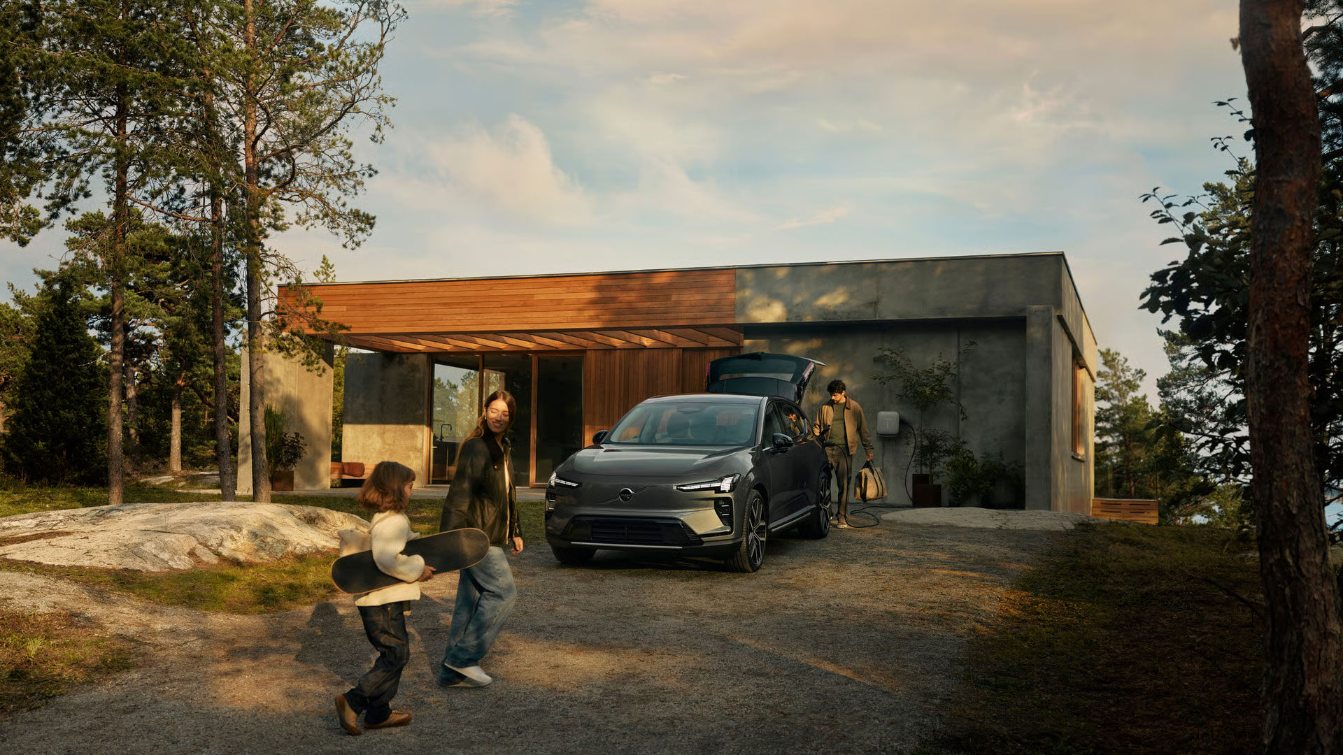 Eine Familie die auf den Volvo EX60 zu geht, der vor einem modernen Haus gepark ist, ein Kind trägt ein Skateboard in der Hand.