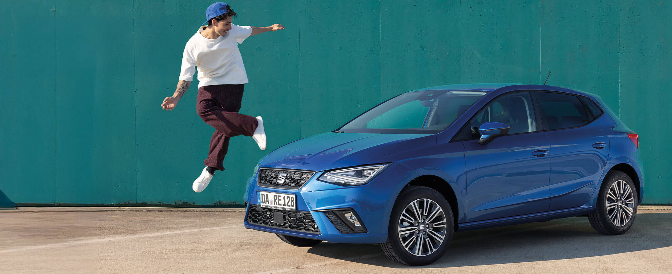 Blauer Seat Ibiza Kompaktwagen, geparkt vor einer grünen Wand. Daneben springt ein junger Mann mit weißem Shirt, blauer Kappe und sportlicher Pose in die Luft.